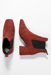 Une paire de bottines en suede couleur rouille avec des talons bloc et des panneaux latéraux en élastique noir, présentées en vues de dessus et de côté sur un fond blanc.