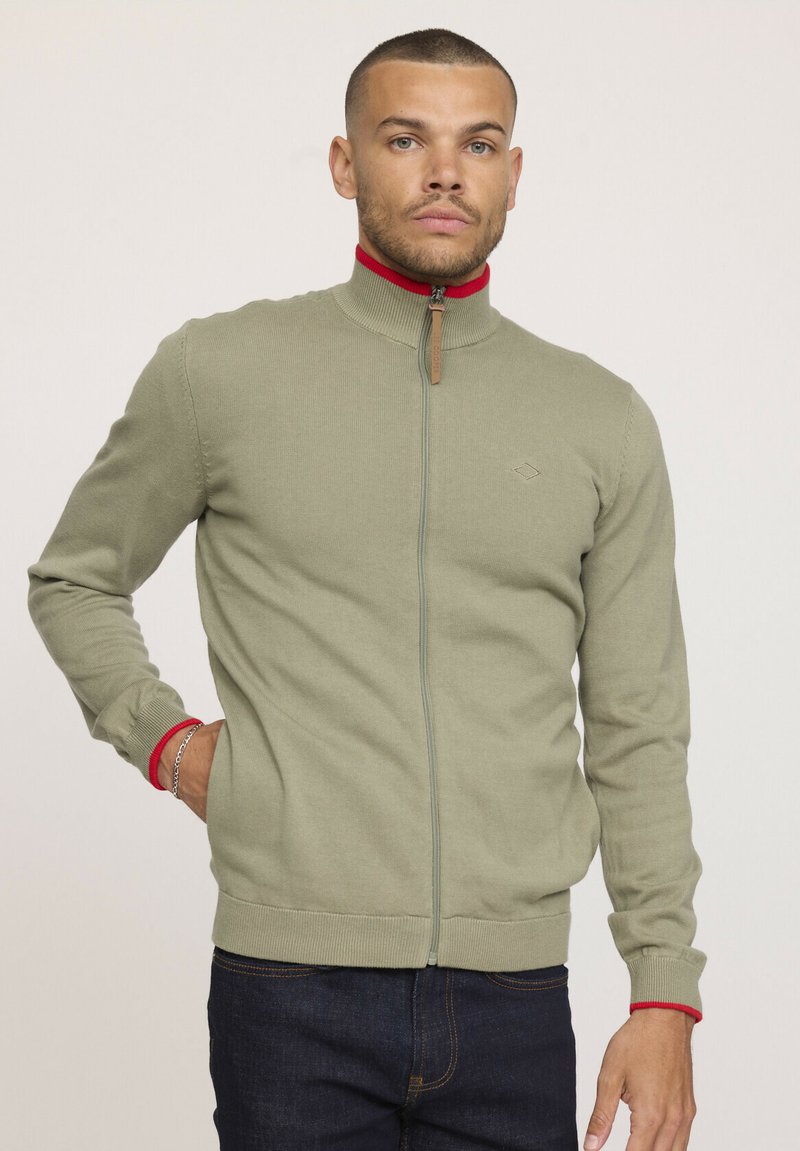 Sweatshirt zippé vert olive clair avec un col montant, accents rouges au col et aux poignets, texture tricotée, et un logo sur la poitrine.