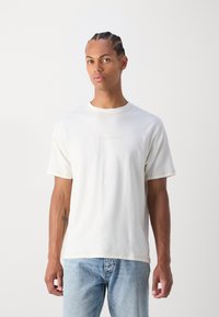 Cremefarvet bomulds t-shirt med korte ærmer, rund hals og kontrastsyning. Har et lille logo trykt på brystet.
