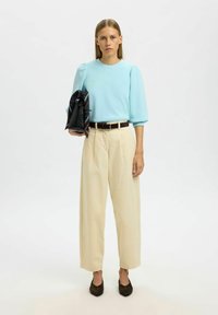 Světle modrý, dlouhý rukáv, texturovaný top spojený s béžovými, vysokými kalhotami s širokými nohavicemi. Hnědý opasek a černé boty. Taška s geometrickým vzorem.