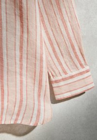 Manche et côté d'une chemise à manches longues rayée beige et rouille avec détail du poignet, affichés sur un fond gris clair.