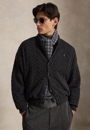 Jeune homme portant une veste en maille torsadée noire, une écharpe à carreaux grise, un pull gris, des lunettes de soleil foncées et un pantalon gris, debout les mains dans les poches.