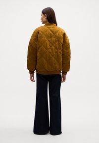 Geknoopte, mosterdgele bomberjack met een geribbelde kraag en manchetten, gecombineerd met een wijde, donkere jeans. De textuur heeft een diamanten patroon.