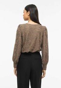 Blouse marron texturée avec motif ondulé, manches longues et col rond, associée à un pantalon noir ajusté. Vue de dos affichée.