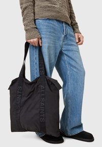 Black nylon tote bag with logo-embossed straps, soft texture, and a relaxed shape, held with one hand, paired with blue jeans.