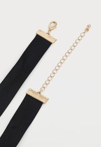 Mustmust kangas kaelakee kuldse tekstuuriga metallkinnitusega ja reguleeritava kuldse ahelaga pikendusega valgel taustal.