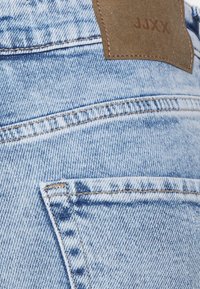 Jeans em denim azul desbotado com um patch em couro castanho rotulado "JUXX." Os recursos incluem costura dupla e um detalhe na parte de trás do bolso.