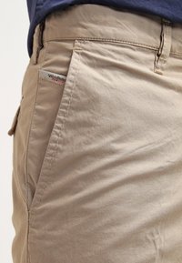 Pantalon beige avec poches latérales visibles et détails cousus, partiellement recouvert par une chemise bleu foncé.