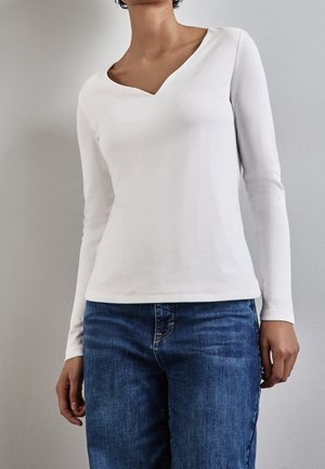 Vrouw draagt een wit langärmelig V-hals topje en een blauwe spijkerbroek, staand tegen een effen lichtgrijze muur.