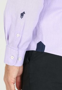 Chemise à manches longues violet clair avec un emblème de cheval sur le poignet, des boutonnages, et des accents latéraux en marine contrastants.