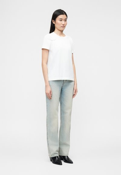 Γυναίκα με μακριά μαύρα μαλλιά, φοράει απλό λευκό κοντομάνικο μπλουζάκι, γαλάζια τζιν κανονική γραμμή και μαύρα μποτάκια με μυτερή μύτη, στέκεται όρθια.