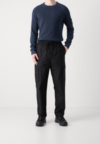 Pull en maille côtelée bleu marine et pantalon cargo noir avec poches latérales. Des baskets noires complètent la tenue, offrant un look décontracté et fonctionnel.