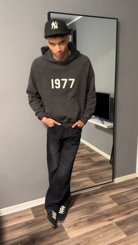 Νέος άνδρας που φοράει μαύρο φούτερ με το «1977», μαύρο παντελόνι και μαύρο καπέλο, στέκεται μέσα κοντά σε έναν μεγάλο καθρέφτη δαπέδου.