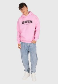 Junger Mann, der einen rosa Kapuzenpullover mit schwarzem Text, eine hellblaue, lässige Jeans und weiße Sneaker trägt. Er steht mit einer Hand in der Tasche.