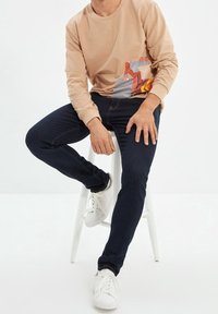 Sudadera de manga larga beige con un estampado geométrico colorido en el frente, combinada con jeans de mezclilla oscura y zapatillas blancas.