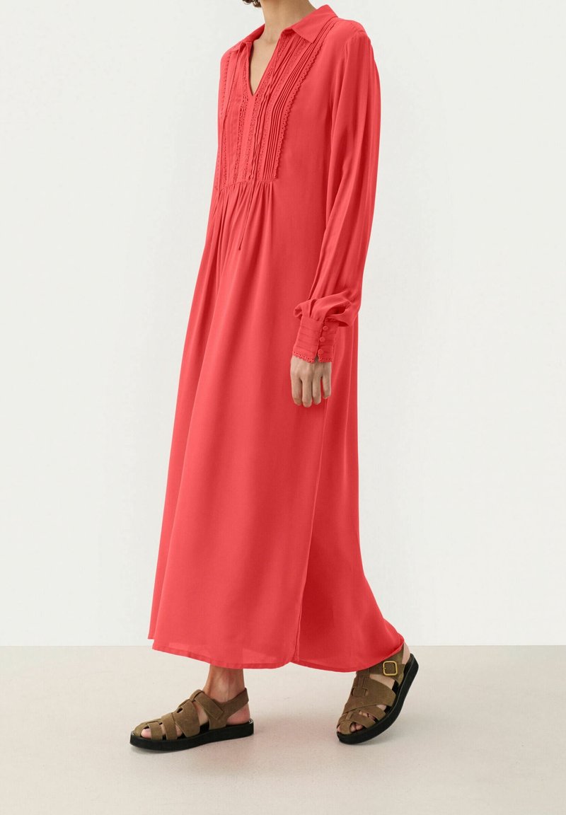 Femme portant une longue robe corail avec des détails en dentelle et des sandales marron à bouts ouverts, debout devant un fond uni blanc cassé.