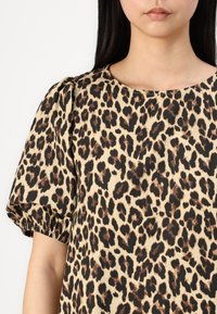Bluză cu imprimeu leopard, mâneci bufante, decolteu rotund, fundal bej și pete negre-maro, realizată dintr-un material neted.