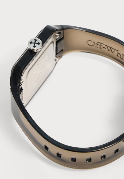 Bracelet de montre noir et beige translucide avec des trous ajustables et une couronne métallique avec un logo à quatre flèches sur le côté du boîtier de la montre.