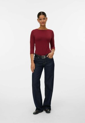 Bordeaux fitted langslieve top gecombineerd met donkerblauwe wijdvallende denim jeans, aangevuld met een zwarte riem met een zilveren gesp. Zwarte platte schoenen.