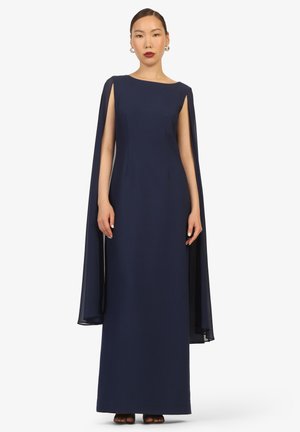 Kobieta w granatowej sukni wieczorowej sięgającej do ziemi z pelerynowymi rękawami, uzupełnionej złotymi kolczykami kołami i ciemnoczerwoną szminką.