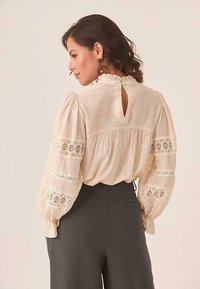 Blusa de crema con mangas abullonadas, acentos de encaje, cierre de ojal en la espalda y tejido texturizado, combinada con pantalones oscuros ajustados.