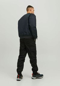 Giacca bomber nera con una texture liscia, abbinata a pantaloni cargo neri. Il modello indossa scarpe sportive con accenti verdi e arancioni.