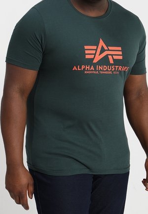 Homme portant un t-shirt vert foncé Alpha Industries avec un logo et un texte orange, associé à un pantalon bleu foncé, debout devant un fond blanc.
