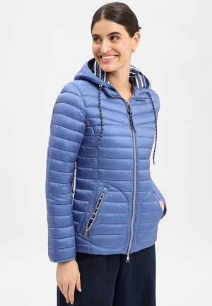 Vrouw die lacht, draagt een blauw doorgestikt gewatteerd jack met capuchon, voorzien van een gestreepte binnenkap en zakken met rits, staand met handen in de zakken.