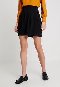 Femme portant une jupe plissée noire, un chemisier à manches longues moutarde, et des mocassins noirs, debout sur un sol blanc devant un fond blanc.