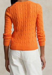 Pull orange en maille torsadée avec manches longues, poignets et ourlet côtelés. Présente un motif texturé ; associé à un pantalon blanc.
