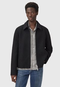 Jonge man in een zwarte ritsjas over een grijs geruit shirt en blauwe jeans, staande met één hand in zijn zak.