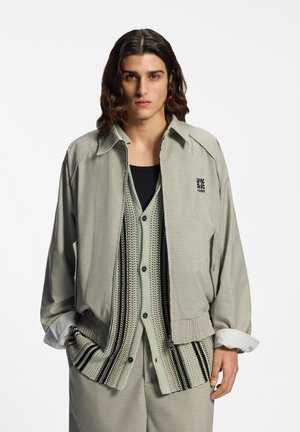 Młody mężczyzna z włosami do ramion, ubrany w jasną kratkowaną kurtkę na szydełkowanej kamizelce zapinanej na guziki i pasujących spodniach, stojący z rękami w kieszeniach.