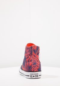 Zapatilla Converse All Star de caña alta con patrón de rayas de tigre en rojo y negro vista desde la parte trasera sobre una superficie y fondo blancos.