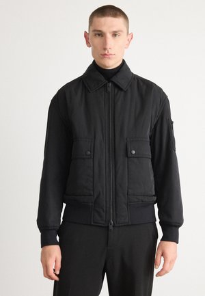 Corta-vento preto com fecho à frente, dois bolsos no peito e punhos canelados. Feito de um tecido suave, apresenta um colarinho dobrável.