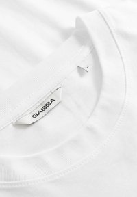 Λευκό T-shirt με στρογγυλή λαιμόκοψη, κοντινή λήψη που δείχνει τον ριμπ ύφασμα στον γιακά και την ραμμένη ετικέτα της μάρκας «GABBA» με την ετικέτα μεγέθους "L" στο εσωτερικό της λαιμόκοψης.
