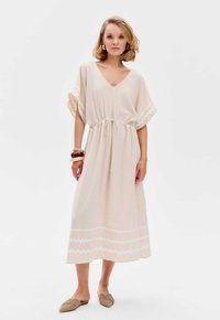 Beige jurk tot de knie met een V-hals, een strikceintuur en vlindermouwen. Kenmerkt zich door witte zigzagpatronen langs de zoom.