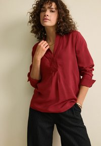 Šarlatová blůza s dlouhým rukávem, V-výstřihem a ohrnutými manžetami, v kombinaci s černými širokými kalhotami s jemným proužkem. Hladká, lehká textilie.