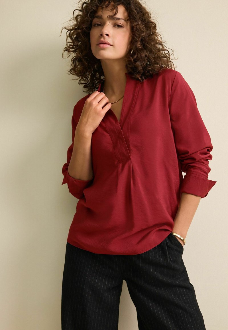 Šarlatová blůza s dlouhým rukávem, V-výstřihem a ohrnutými manžetami, v kombinaci s černými širokými kalhotami s jemným proužkem. Hladká, lehká textilie.