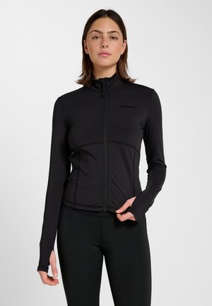 Jonge vrouw die een zwarte sportjas met volledige ritssluiting en zwarte legging draagt, staand tegen een effen witte achtergrond.