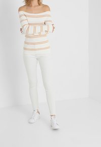 Woman standing with arms crossed wearing off-shoulder white sweater with orange stripes, white skinny jeans, and white sneakers.