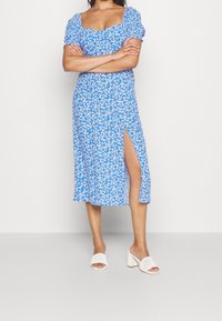 Blaues Blumenkleid mit Puffärmeln, quadratischem Ausschnitt und seitlichem Schlitz. Aus leichtem Stoff gefertigt. Getragen mit weißen Blockabsatzsandalen.