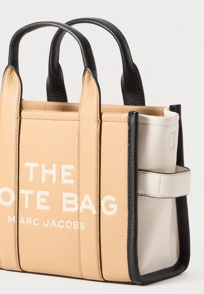 Mini tote soma bēšā un melnā ādā ar izteiktu iegravētu tekstu "TOTE SOMA" un "MARC JACOBS"; aprīkota ar divām augšējām rokturiem un sānu kabatu.