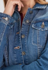 Kurtka jeansowa w średnim odcieniu niebieskiego z metalowymi guzikami, przetarciami i dwoma kieszeniami na piersi. Bawełniany materiał o klasycznym kroju.
