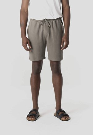 Hombre con pantalones cortos texturizados color topo con cordón, camiseta blanca y sandalias negras de doble tirante, de pie sobre fondo blanco.