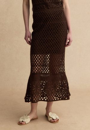 Unterkörper einer Person, die einen braunen gehäkelten Midirock mit Blumenmustern und beige Slipper mit Blumendekorationen trägt.