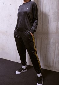 Osoba stojí s rukama v kapsách, oblékající černý sametový sportovní komplet se zlatými pruhy na kalhotách a černo-bílé tenisky Adidas.