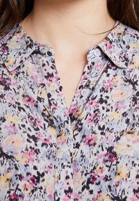 Blus med blommönster i lätt tyg; har en krage, knappstängning och en blandning av rosa, gult och svart.