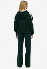 Dunkelgrüner Kapuzenpullover mit weißen Seitenstreifen, kombiniert mit passenden Schlaghosen. Weicher Stoff, lässiges Design und lockere Passform.