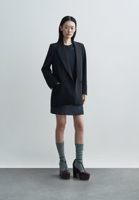 Czarna, luźna marynarka z rozciętym kołnierzem, zestawiona z ciemną dżinsową spódnicą oraz szarymi prążkowanymi skarpetami. Stylizację dopełniają buty na platformie.