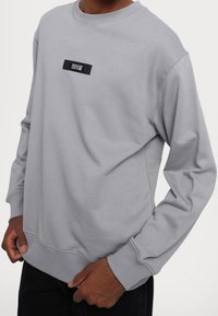 Grå sweatshirt med rund halsringning, ribbstickade ärmmuddar och en liten svart etikett på bröstet. Tillverkad av ett mjukt, strukturerat tyg.